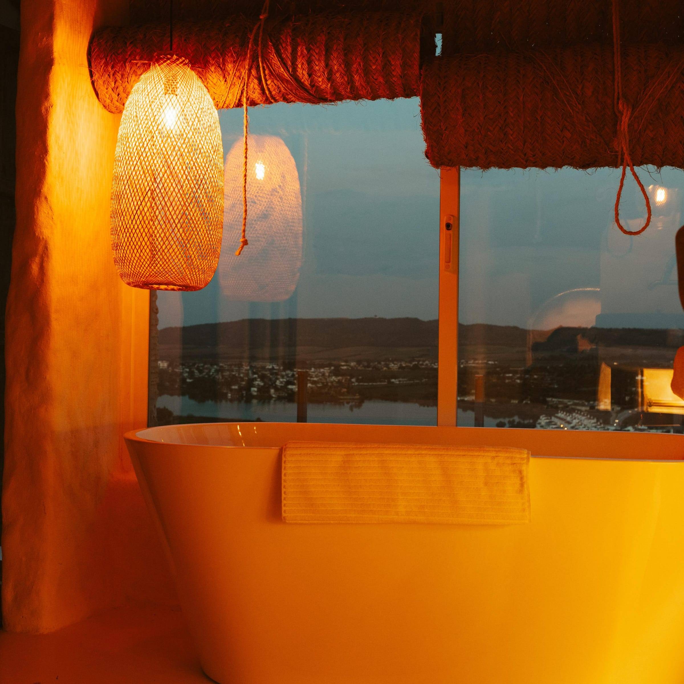 Bañera exenta frente a un ventanal con vistas al lago al atardecer, a la luz de una lámpara de fibra natural