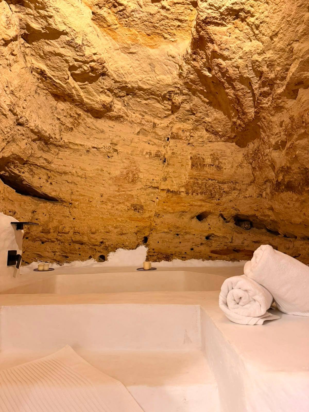 Bañera excavada en el suelo bajo el techo de roca, con toallas blancas enrolladas en el borde