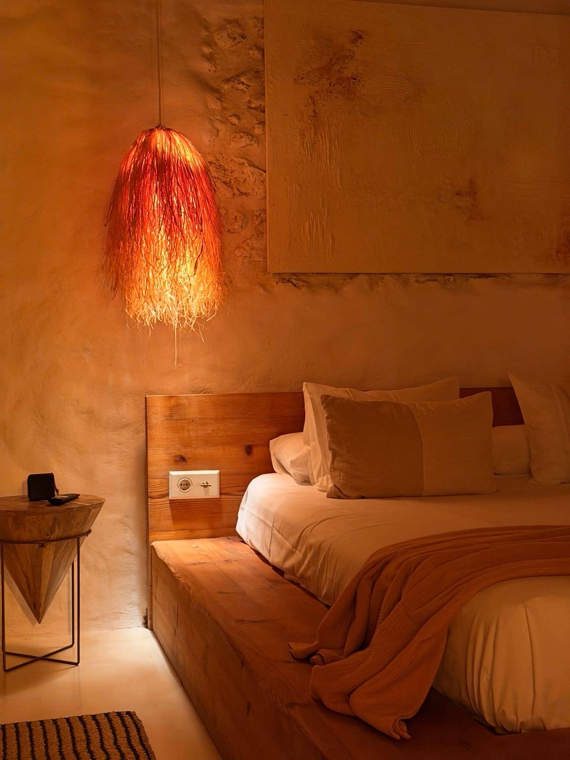 Lámpara de fibra natural encendida sobre una cama de madera con ropa de tonos tierra