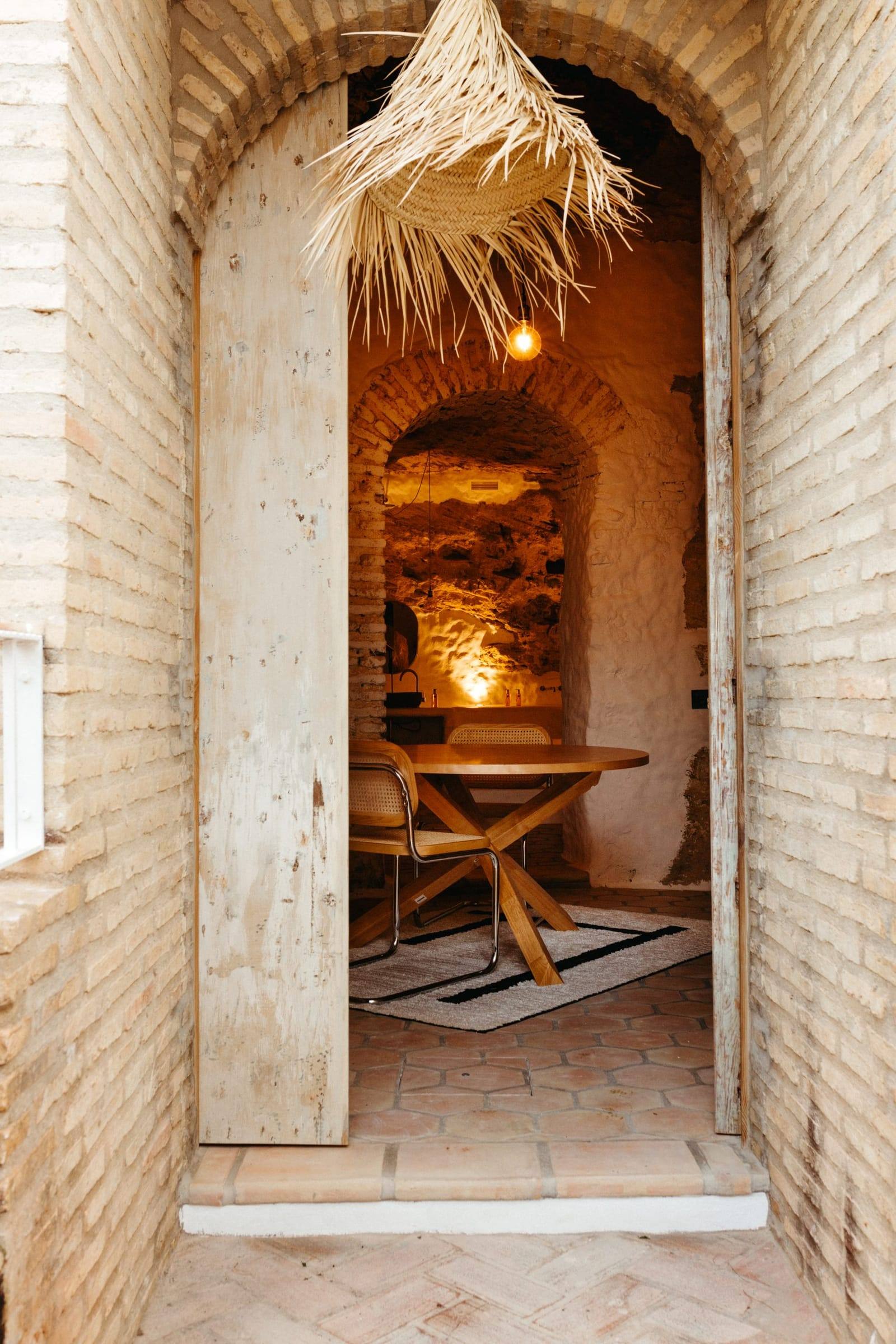 Entrada por una puerta de madera bajo un arco de ladrillo, con el comedor iluminado al fondo