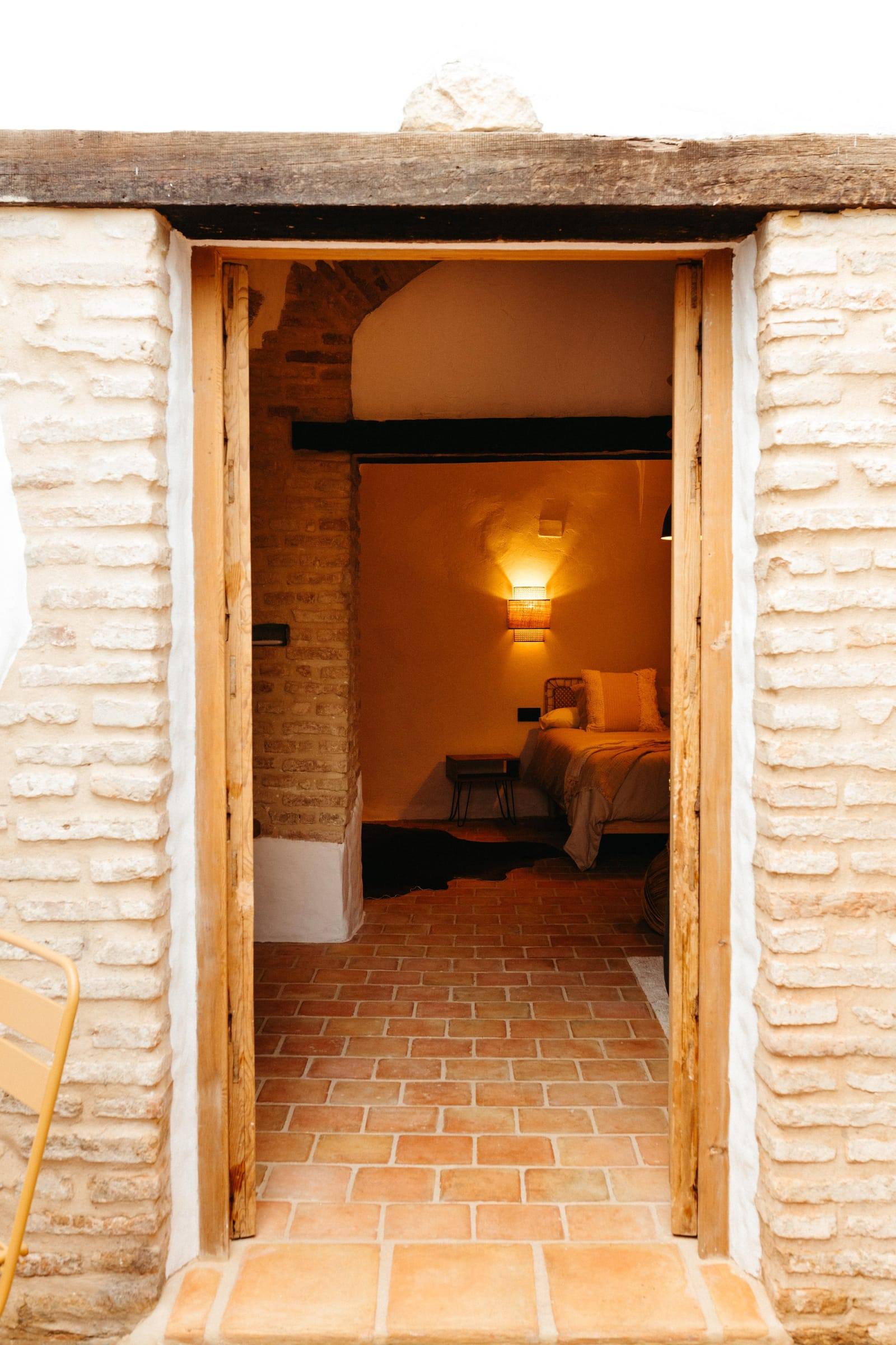 Puerta de madera abierta en un marco de ladrillo que deja ver el dormitorio iluminado, sobre suelo de barro