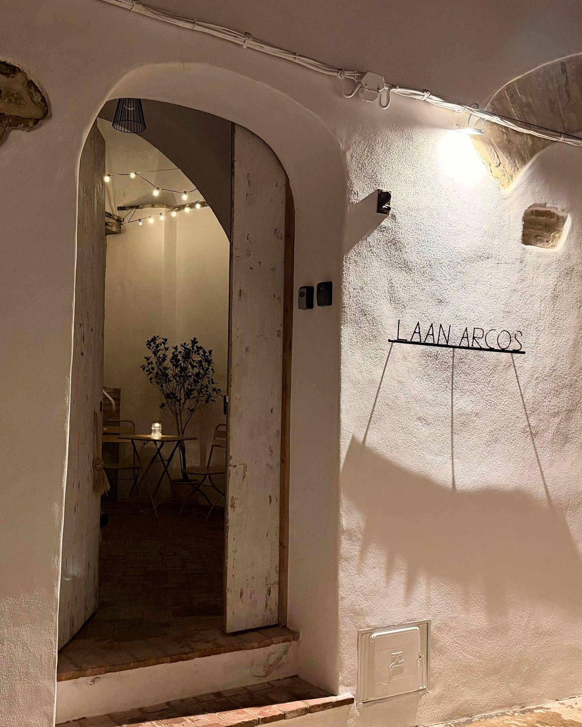 Puerta de Laan Arcos abierta de noche bajo un arco encalado, con el patio iluminado por guirnaldas al fondo y el nombre en letras de metal en la fachada