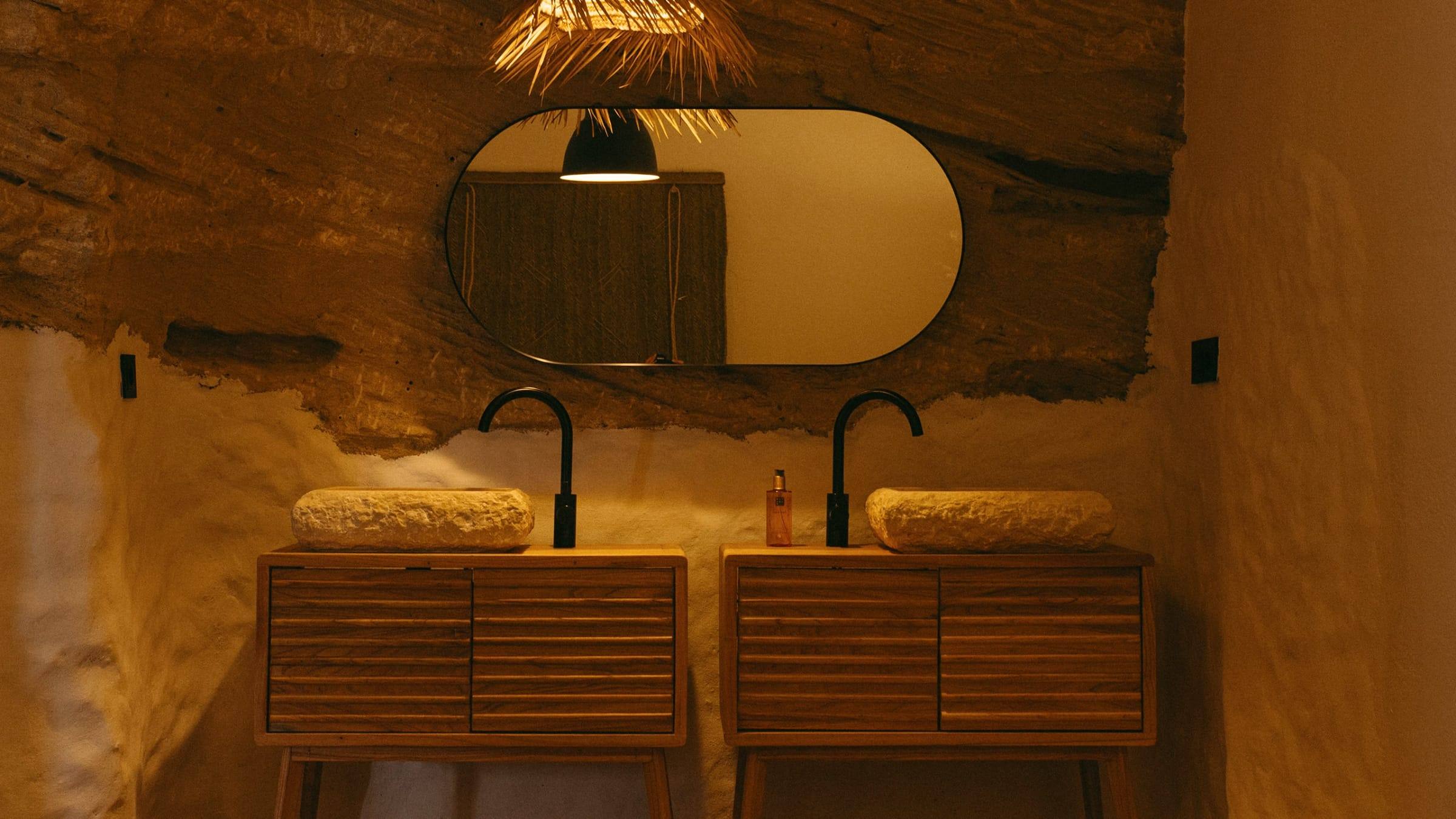 Baño de Verdolaga bajo la roca, con dos lavabos de piedra sobre muebles de madera, grifos negros y un espejo ovalado