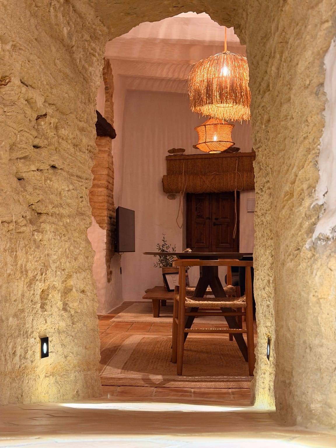 Paso excavado en la piedra que lleva a un salón con mesa de madera y lámparas de fibra
