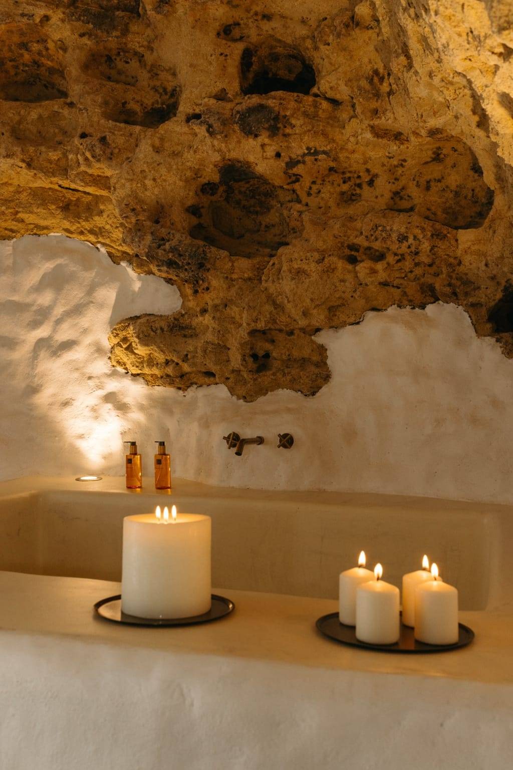 Velas encendidas en el borde de una bañera encalada, bajo una pared de roca natural