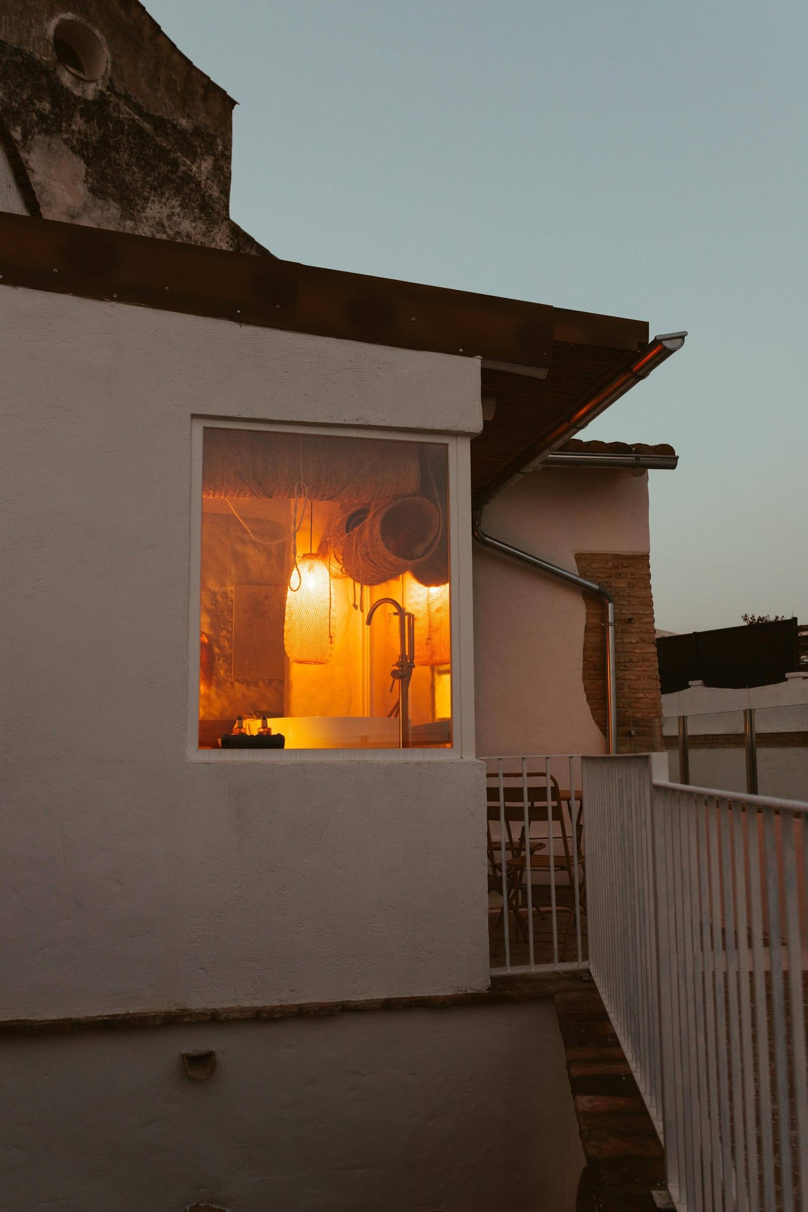 Ventana iluminada de la estancia vista desde la terraza al anochecer, junto a una barandilla blanca